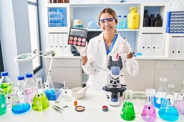 Fototapeta premium Young blonde woman working at scientist laboratory with make up smiling happy and positive, thumb up doing excellent and approval sign