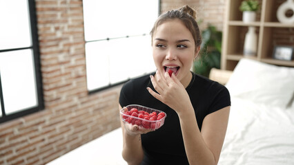 Young beautiful hispanic woman eating raspberries sitting on bed at bedroom