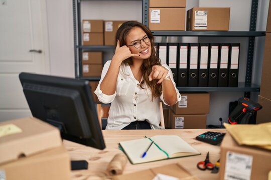 Young hispanic woman working at small business ecommerce smiling doing talking on the telephone gesture and pointing to you. call me.