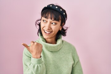 Young beautiful woman standing over pink background smiling with happy face looking and pointing to the side with thumb up.