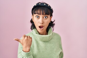 Young beautiful woman standing over pink background surprised pointing with hand finger to the side, open mouth amazed expression.