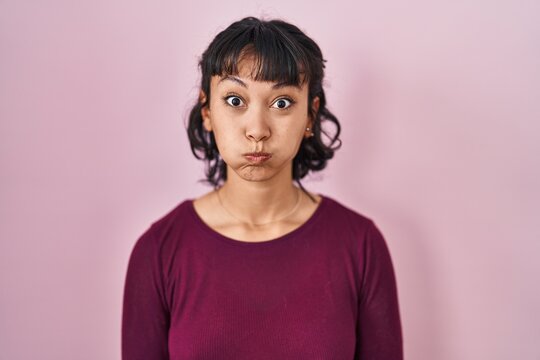 Young beautiful woman standing over pink background puffing cheeks with funny face. mouth inflated with air, crazy expression.