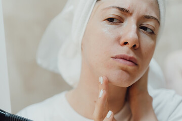 After the shower, the body and head wrapped in a towel. A young woman looks at her face in the mirror and thinks about how to take care of her facial skin. Pimples and acne.