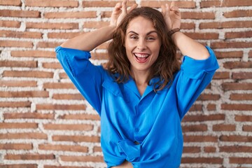 Beautiful brunette woman standing over bricks wall posing funny and crazy with fingers on head as bunny ears, smiling cheerful