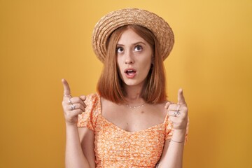 Young redhead woman standing over yellow background wearing summer hat amazed and surprised looking...