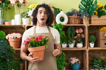 Hispanic man with curly hair working at florist shop holding plant scared and amazed with open mouth for surprise, disbelief face