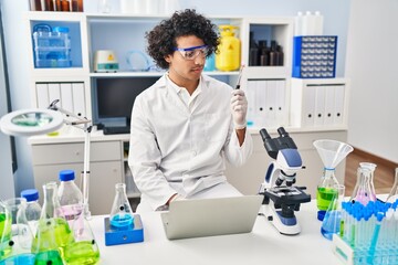 Young hispanic man wearing scientist uniform using laptop analysing blood test tube at laboratory