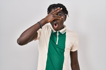 African man with dreadlocks standing over isolated background peeking in shock covering face and...