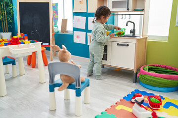 Adorable hispanic toddler playing with play kitchen standing at kindergarten