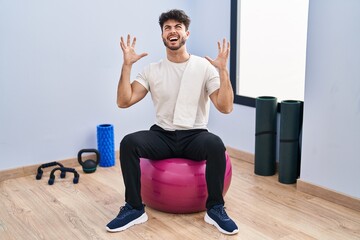 Hispanic man with beard sitting on pilate balls at yoga room crazy and mad shouting and yelling with aggressive expression and arms raised. frustration concept.