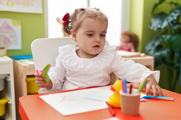Adorable blonde toddler student sitting on table holding scissors at kindergarten