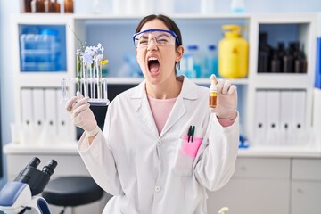 Young brunette woman working at scientist laboratory angry and mad screaming frustrated and furious, shouting with anger. rage and aggressive concept.