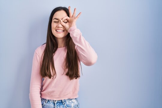 Young brunette woman standing over blue background doing ok gesture with hand smiling, eye looking through fingers with happy face.