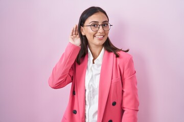 Young hispanic woman wearing business clothes and glasses smiling with hand over ear listening an hearing to rumor or gossip. deafness concept.