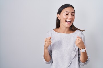 Young hispanic woman standing over white background very happy and excited doing winner gesture with arms raised, smiling and screaming for success. celebration concept.