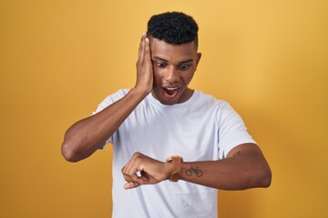 Young hispanic man standing over yellow background looking at the watch time worried, afraid of...