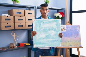 Young hispanic man holding canvas at artist studio in shock face, looking skeptical and sarcastic,...