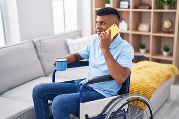 Young latin man talking on smartphone drinking coffee sitting on wheelchair at home