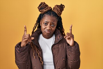 African woman with braided hair standing over yellow background pointing up looking sad and upset,...