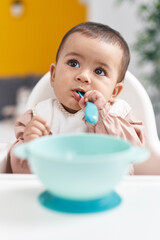 Adorable hispanic baby sitting on highchair sucking spoon at home