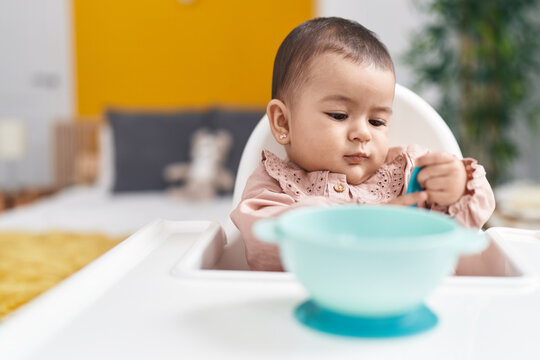 Adorable hispanic baby sitting on highchair holding spoon at bedroom