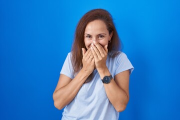 Brunette woman standing over blue background laughing and embarrassed giggle covering mouth with...