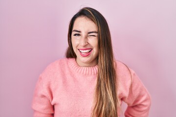 Young hispanic woman standing over pink background winking looking at the camera with sexy...