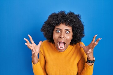 Black woman with curly hair standing over blue background crazy and mad shouting and yelling with aggressive expression and arms raised. frustration concept.