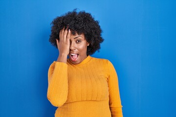 Fototapeta na wymiar Black woman with curly hair standing over blue background covering one eye with hand, confident smile on face and surprise emotion.