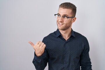 Young caucasian man standing over isolated background smiling with happy face looking and pointing to the side with thumb up.