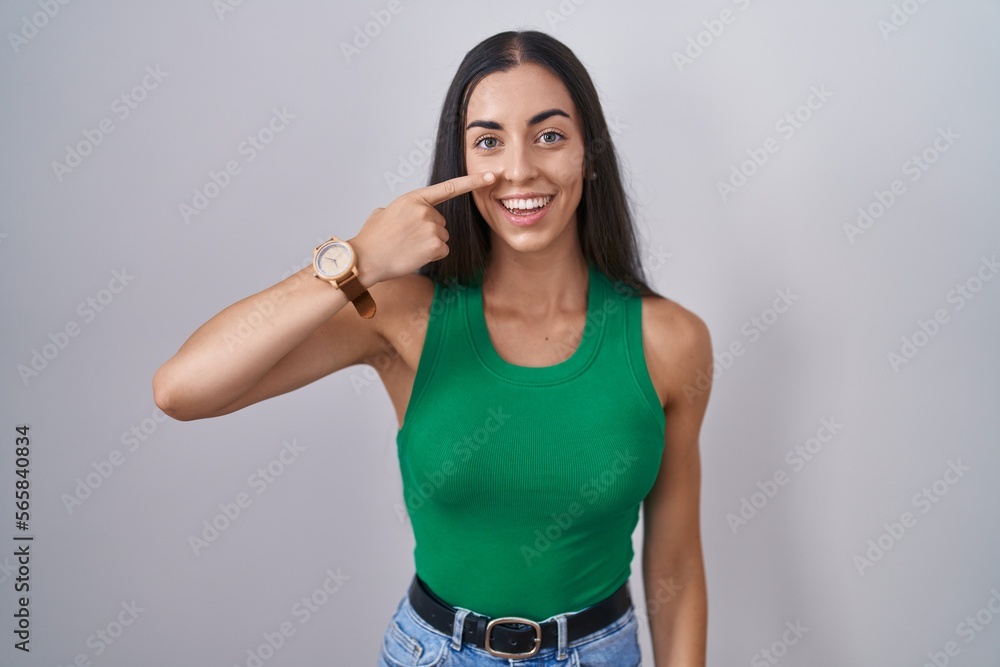 Poster young woman standing over isolated background pointing with hand finger to face and nose, smiling ch