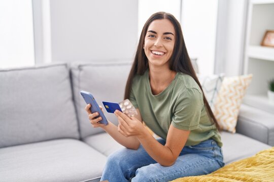 Young beautiful hispanic woman using smartphone and credit card holding pills at home