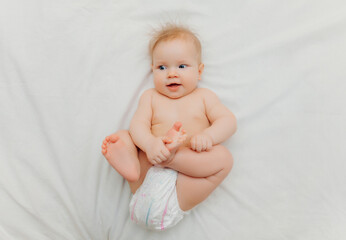 a happy joyful baby in diapers is lying on a white bed and holding his leg. High-quality photo. place for text