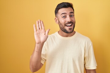 Handsome hispanic man standing over yellow background waiving saying hello happy and smiling, friendly welcome gesture