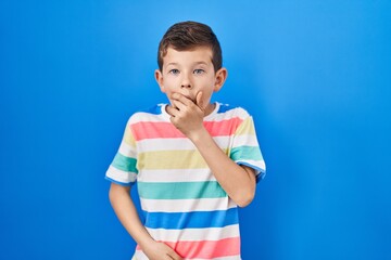 Young caucasian kid standing over blue background looking fascinated with disbelief, surprise and amazed expression with hands on chin