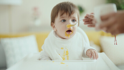 Fototapeta premium Adorable hispanic toddler sitting on highchair eating food at home