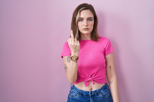 Blonde caucasian woman standing over pink background showing middle finger, impolite and rude fuck off expression