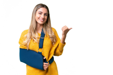 Young Uruguayan woman with broken arm and wearing a sling over isolated background pointing to the side to present a product
