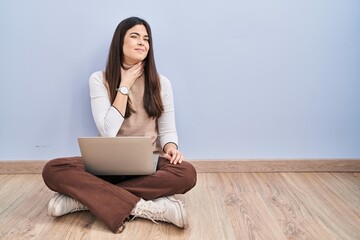 Young brunette woman working using computer laptop sitting on the floor touching painful neck, sore throat for flu, clod and infection