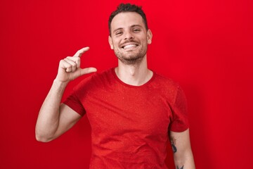 Fototapeta premium Young hispanic man standing over red background smiling and confident gesturing with hand doing small size sign with fingers looking and the camera. measure concept.
