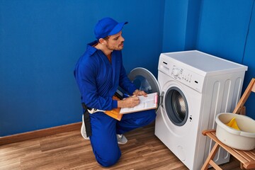 Young hispanic man technician repairing washing machine writing on document at laundry room