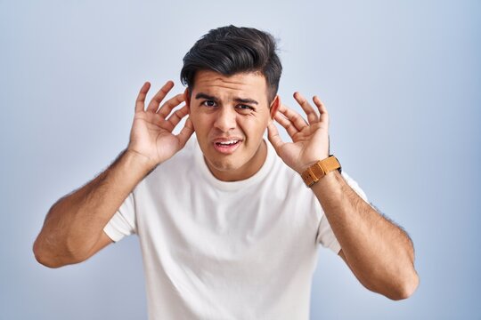 Hispanic man standing over blue background trying to hear both hands on ear gesture, curious for gossip. hearing problem, deaf