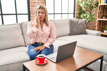 Young hispanic woman using laptop sitting on the sofa at home looking stressed and nervous with hands on mouth biting nails. anxiety problem.