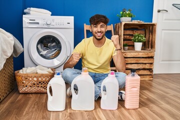 Arab man with beard doing laundry sitting on the floor with detergent bottle celebrating surprised and amazed for success with arms raised and eyes closed