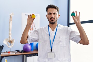 Young hispanic physiotherapist man holding strength balls to train hand muscles skeptic and...
