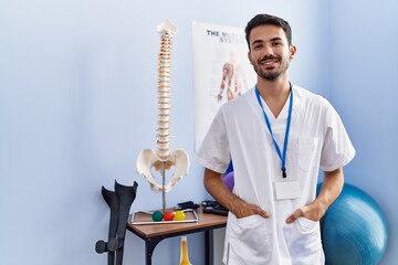 Young hispanic man wearing physiotherapist uniform standing at rehab clinic