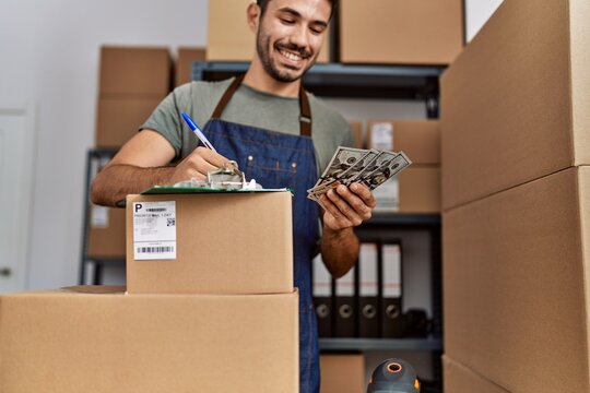 Young hispanic man business worker writing on clipboard holding dollars at storehouse