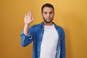 Hispanic man standing over yellow background doing stop sing with palm of the hand. warning...