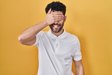 Arab man standing over yellow background smiling and laughing with hand on face covering eyes for surprise. blind concept.