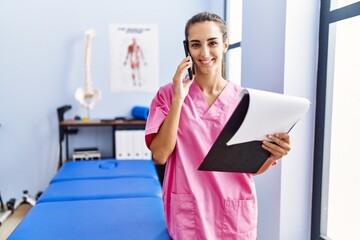 Young hispanic woman wearing physiotherapist uniform talking on the smartphone at physiotherpy clinic